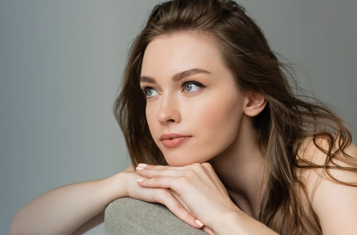 Woman with brown hair resting chin on hand, looking thoughtfully to the side against gray background