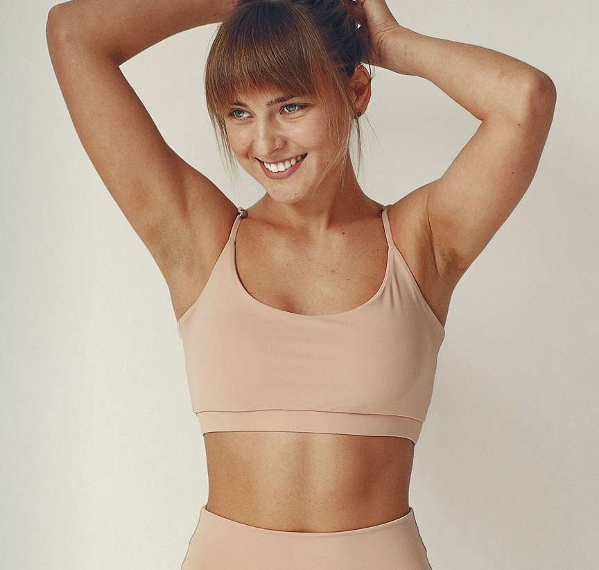 Woman in a beige sports bra smiling with hands behind her head