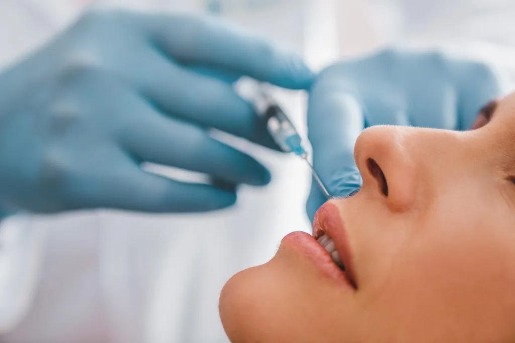 Practitioner in blue gloves injecting filler into patient's lips in profile