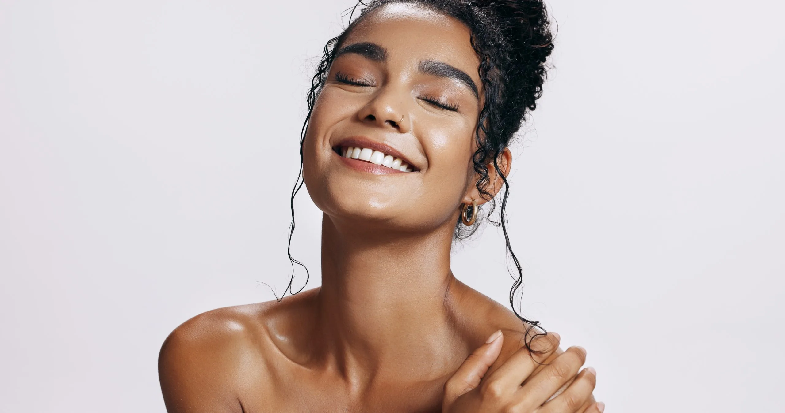 Smiling person with glowing skin touching their shoulder against a neutral background
