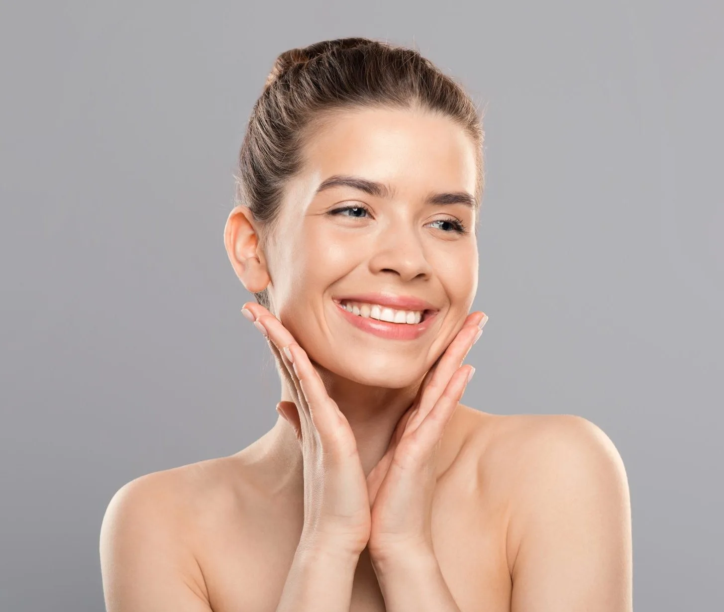 Woman with facial mask smiling and touching her cheeks against gray background