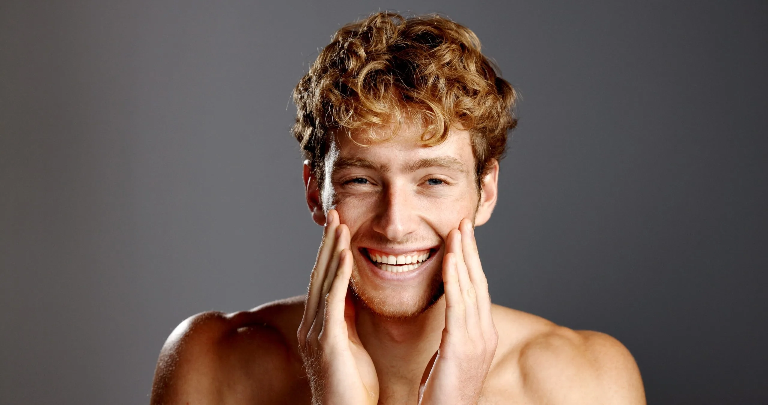 Young man with curly hair smiling while touching his face